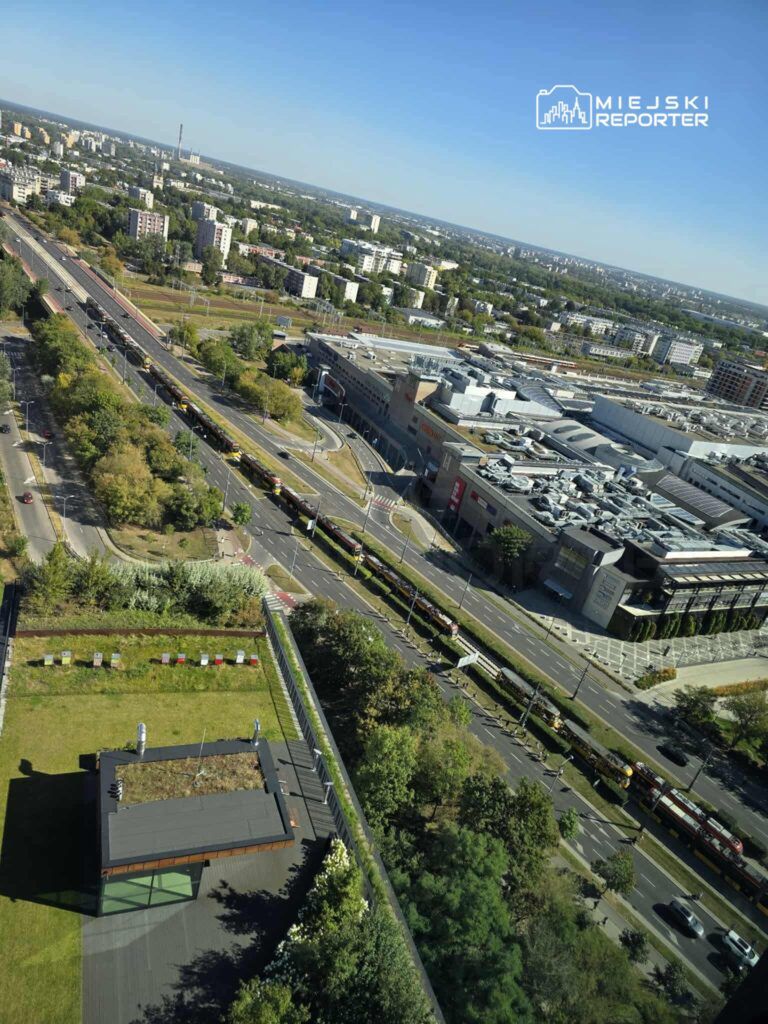 Widok na nowoczesny biurowiec z przeszkloną fasadą i zieleń w okolicy, z ruchliwą ulicą oraz tramwajami w tle.