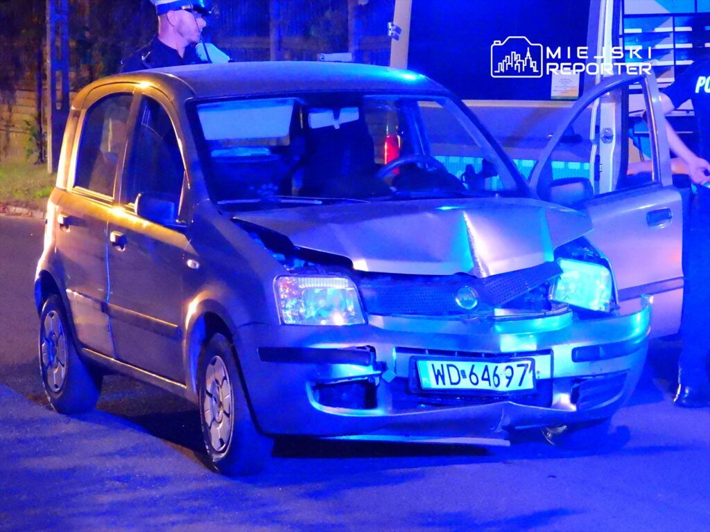 Uszkodzony samochód osobowy zaparkowany na ulicy, w tle policjant z notatnikiem.