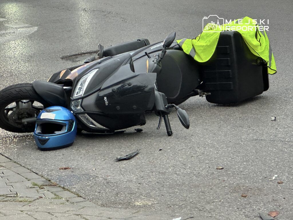 Skuter leży na boku na jezdni, obok niego niebieski kask oraz żółta kamizelka odblaskowa.