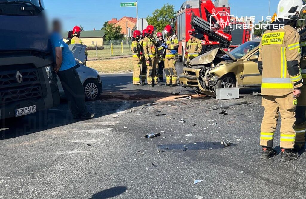 Grupa strażaków w mundurach omawia sytuację przy zderzeniu dwóch pojazdów na drodze, wokół leżą odłamki.