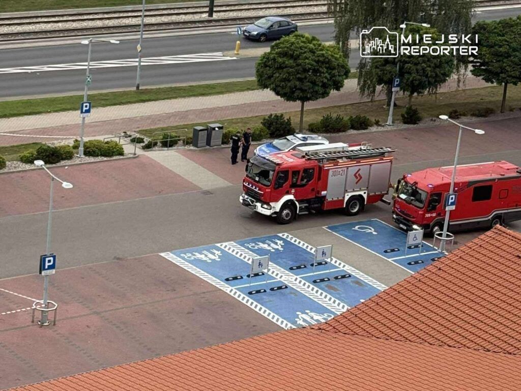 Dwa pojazdy straży pożarnej zaparkowane na parkingu, obok grupy osób rozmawiających w pobliżu.