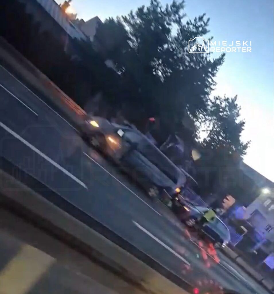 Samochód osobowy zatrzymany na drodze, obok stoi policjant w kamizelce odblaskowej, w tle widoczne drzewa i budynki.