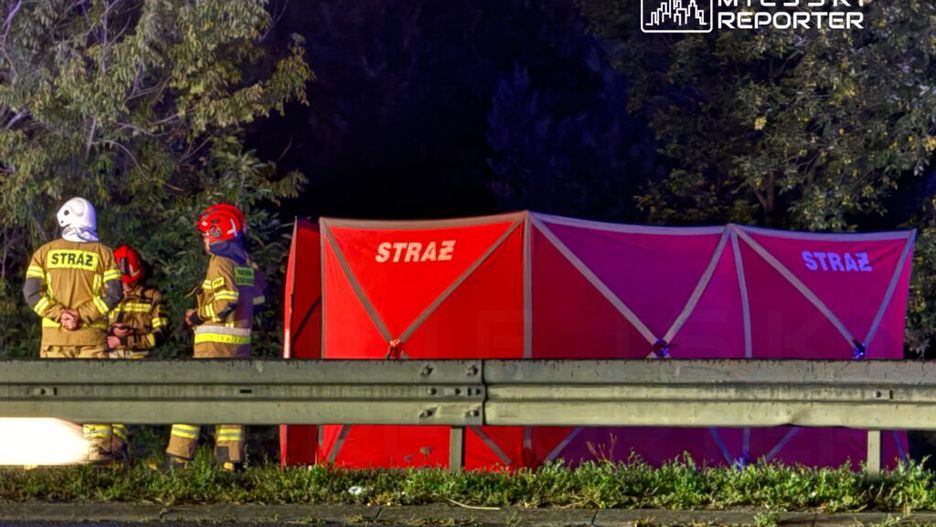 Grupa strażaków w mundurach rozmawia obok czerwonego namiotu z napisem "STRAŻ" przy drodze.