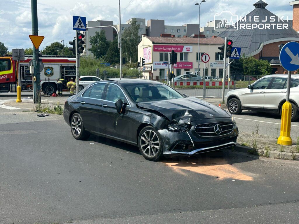 W zdarzeniu drogowym na skrzyżowaniu ulic doszło do kolizji dwóch samochodów. Na miejscu interweniują służby ratunkowe, w tym straż pożarna. Uszkodzony pojazd to czarny Mercedes z widocznymi zniszczeniami. Ruch w okolicy jest wstrzymany.