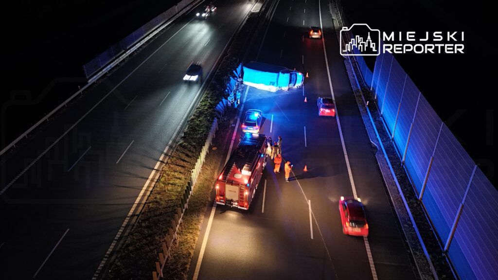 Na drodze leży przewrócony niebieski pojazd, a obok pracują strażacy w pomarańczowych strojach.