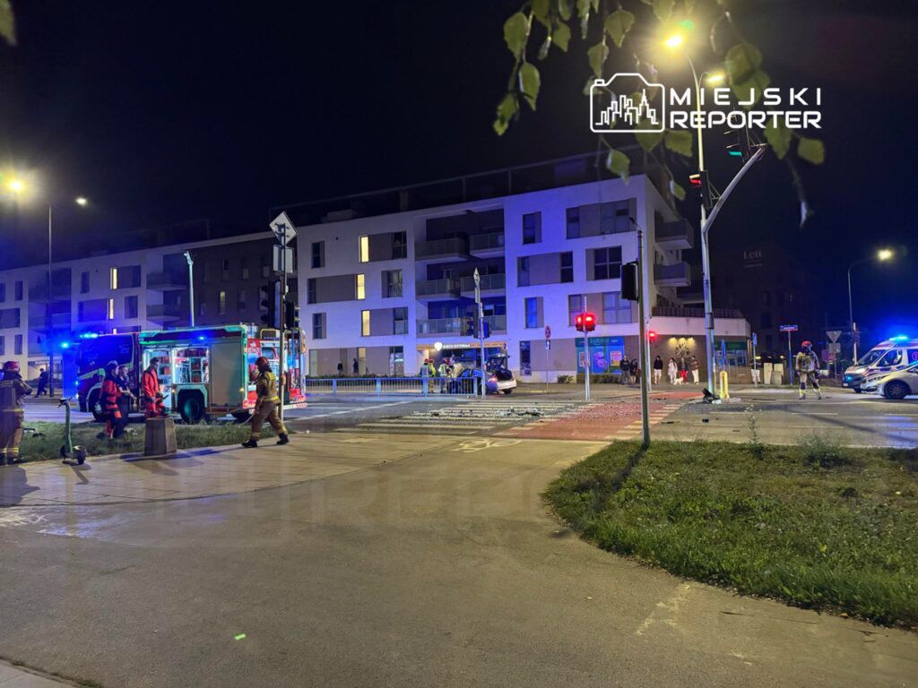 Grupa strażaków i ratowników medycznych pracuje przy wypadku drogowym w nocy, obok nowoczesnych budynków mieszkalnych.