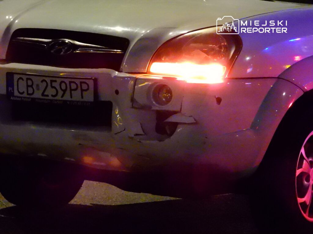 Biały samochód z uszkodzoną tylną częścią, zaparkowany na ulicy, z włączonymi światłami.