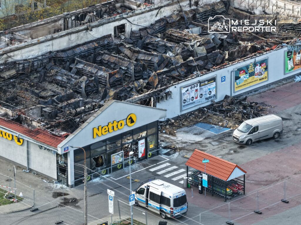 Zniszczony budynek sklepu Netto z wypaloną konstrukcją i uszkodzonymi szybami, w otoczeniu zaparkowanych pojazdów.