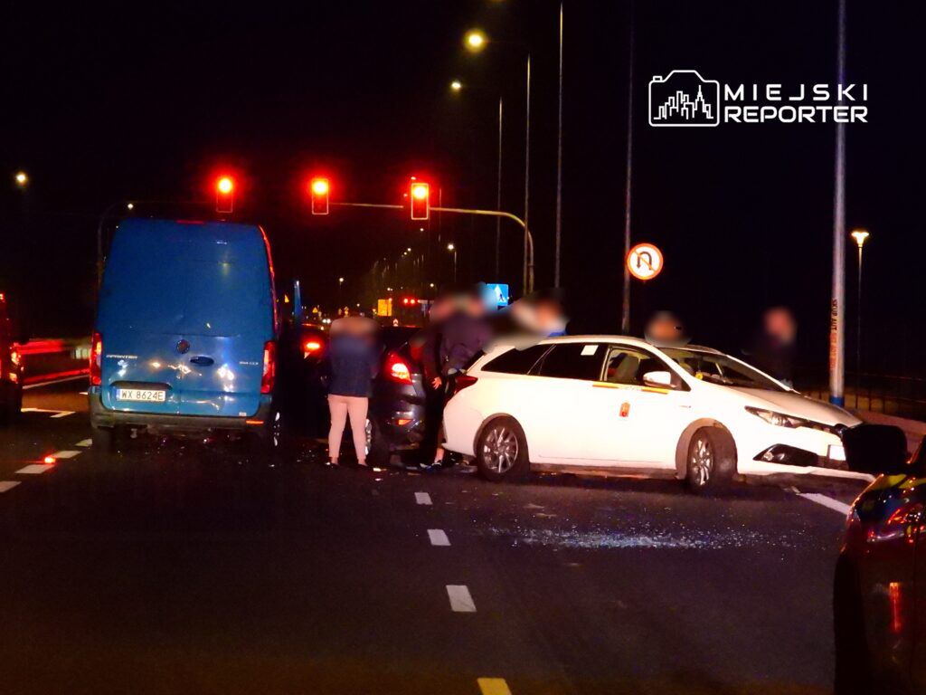 Trzy samochody, w tym biały pojazd i niebieski furgon, zderzone na jezdni w nocy, otoczone przez kilka osób.