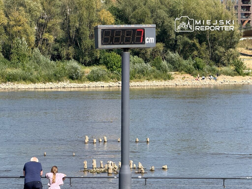 Dziecko w różowej koszulce i starszy mężczyzna obserwują rzekę, obok wyświetlacz z wysokością wody 68,7 cm.