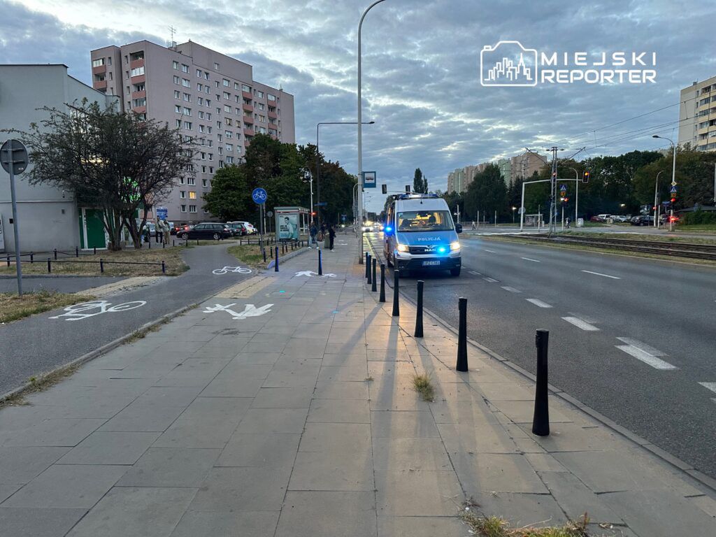 Policyjny radiowóz z włączonymi sygnałami świetlnymi zatrzymany na ulicy w pobliżu przystanku autobusowego.