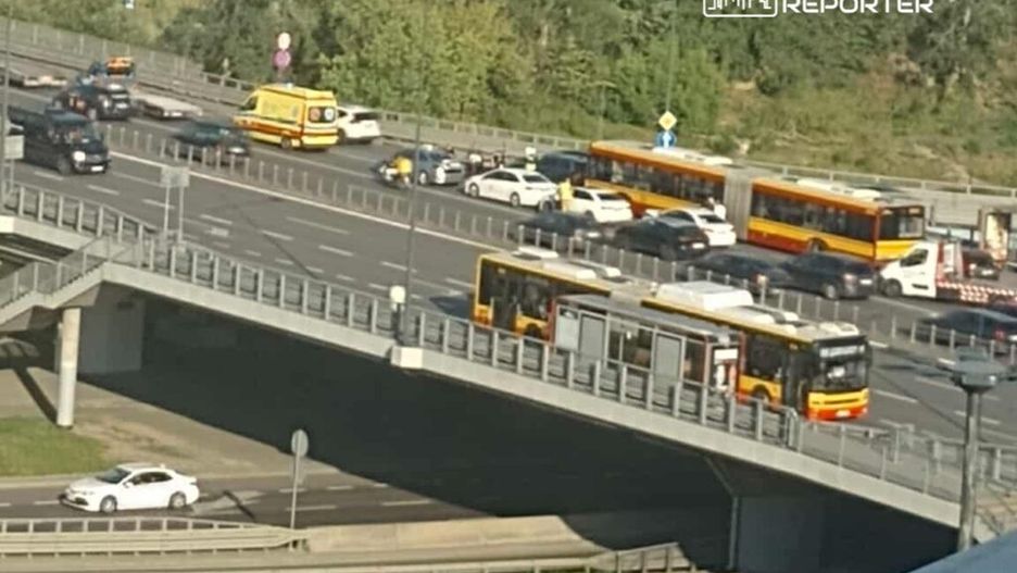 Zatłoczona ulica w mieście, na pierwszym planie widoczne autobusy miejskie oraz samochody osobowe. W tle widać zieleń i znaki drogowe. Obraz przedstawia codzienny ruch komunikacyjny oraz infrastrukturę transportową w urbanistycznym otoczeniu.