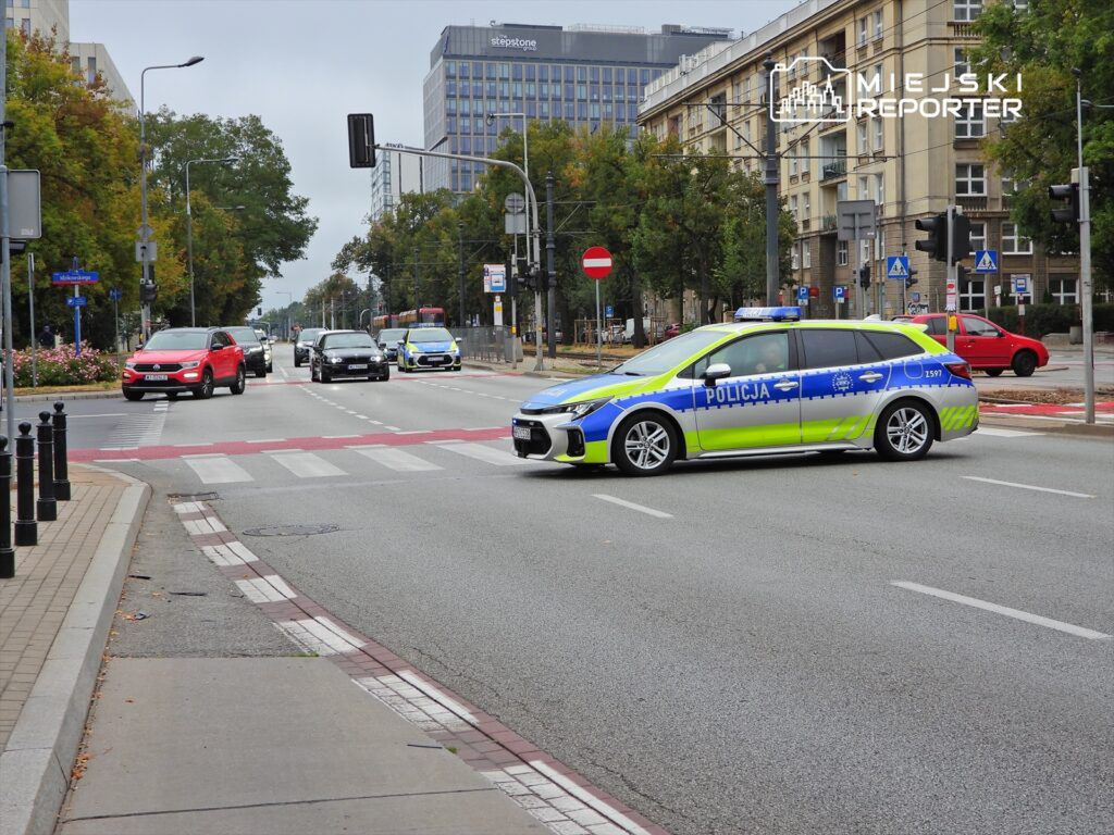 Radiowóz policyjny zatrzymany na skrzyżowaniu, obok poruszające się samochody osobowe w miejskim otoczeniu.