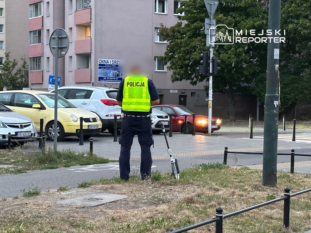 Policjant w kamizelce odblaskowej kieruje ruchem na skrzyżowaniu obok parkingu z zaparkowanymi samochodami.
