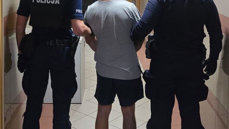 Mężczyzna prowadzony przez dwóch policjantów w korytarzu budynku.