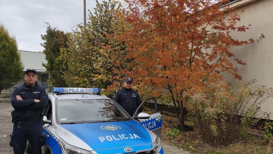 Dwaj policjanci stoją obok nieoznakowanego radiowozu w otoczeniu drzew w jesiennych kolorach.