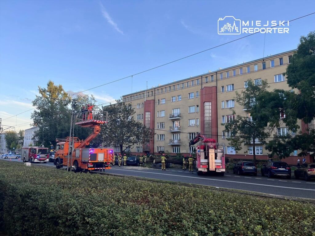 Strażacy w hełmach pracują przy wozie strażackim, a podnośnik gaśniczy sięga do okna budynku mieszkalnego.