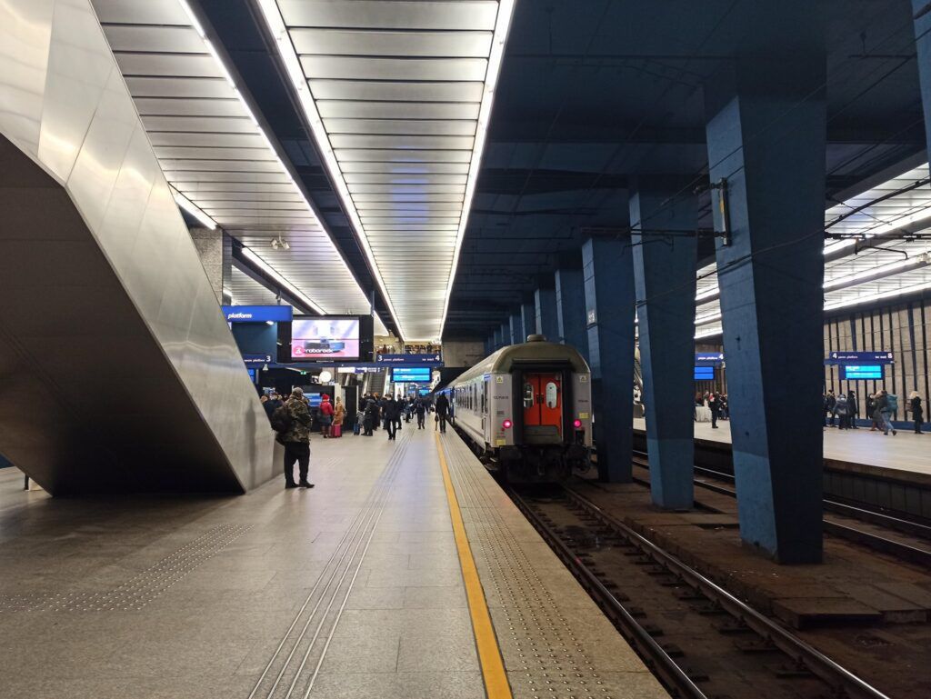 Pasażerowie czekają na peronie stacji kolejowej, obok zatrzymany pociąg w nowoczesnym wnętrzu z niebieskimi filarami.