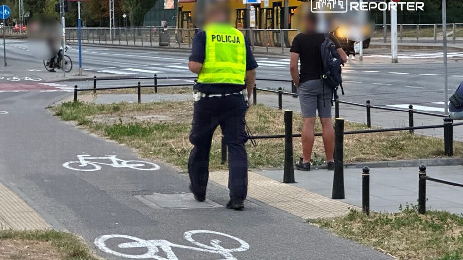 Policjant w kamizelce odblaskowej rozmawia z mężczyzną stojącym obok ścieżki rowerowej w miejskim otoczeniu.