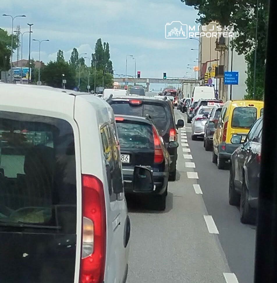 Korki na ulicy w mieście, z widokiem na zator drogowy. Ruch samochodowy wstrzymany, a pojazdy stoją w kolejce. W tle sygnalizacja świetlna oraz znaki drogowe. Obraz ilustruje problemy komunikacyjne w godzinach szczytu.