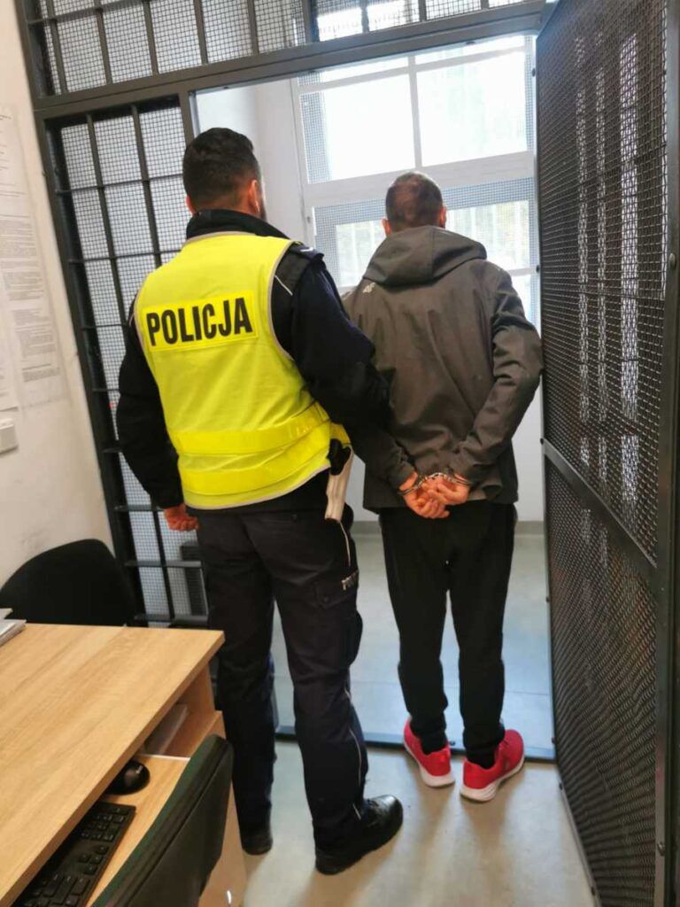 Mężczyzna w szarej bluzie z rękami skrępowanymi za plecami prowadzony przez policjanta w kamizelce.