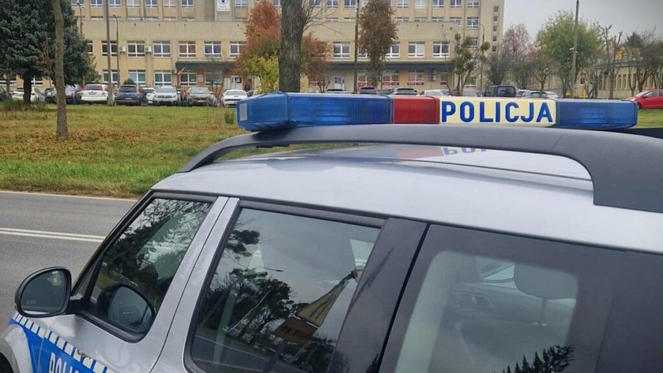 Radiowóz policyjny zatrzymany przy szpitalu, z widocznymi niebieskimi sygnalizatorami na dachu.
