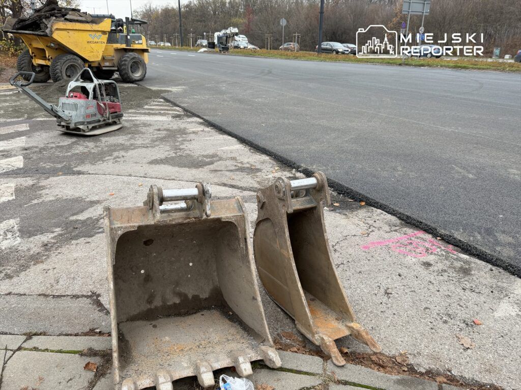 Na drodze leżą dwie łyżki od koparki, a w tle widać pojazd budowlany transportujący asfalt.