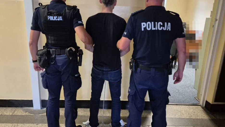 Mężczyzna w kajdankach prowadzony przez dwóch policjantów w budynku z jasnymi ścianami.