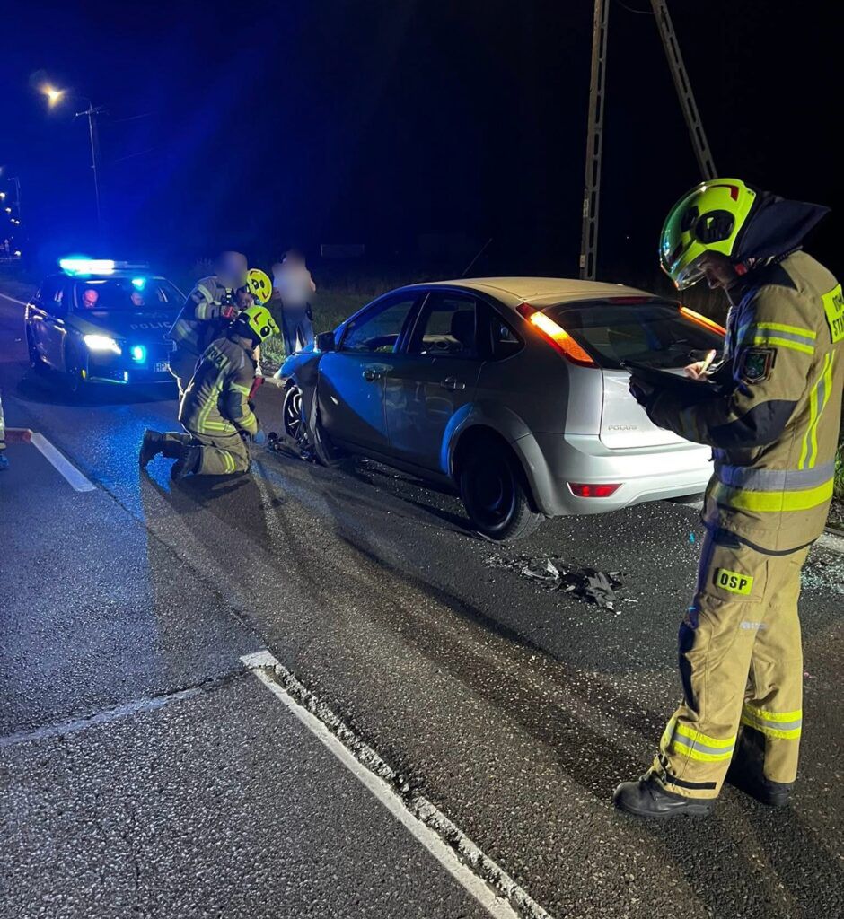 Strażacy w mundurach na drodze, oceniają uszkodzenia samochodu po kolizji, w tle policyjny radiowóz z włączonymi światłami.