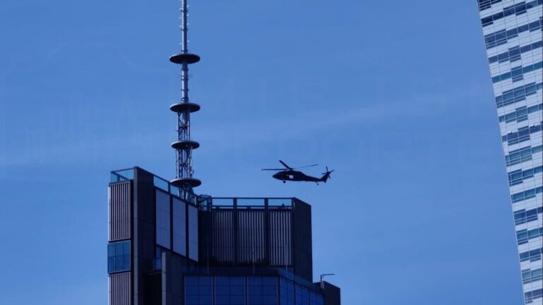 Czarny helikopter przelatuje obok nowoczesnego biurowca z przeszkloną fasadą i anteną na dachu.