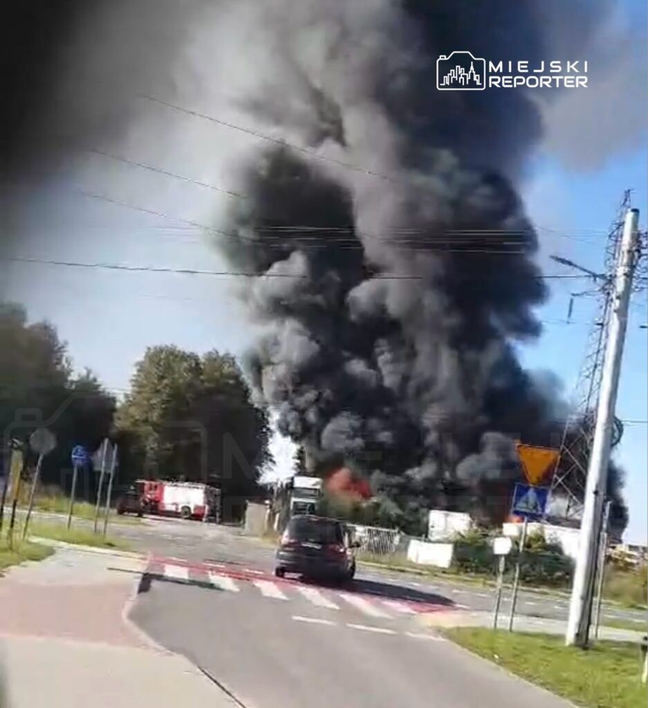 Gęsty czarny dym unoszący się z miejsca pożaru w pobliżu drogi, w tle widoczny wóz strażacki.