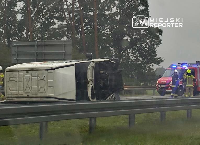 Przewrócony na bok samochód dostawczy na drodze, obok pracują strażacy i karetka pogotowia.