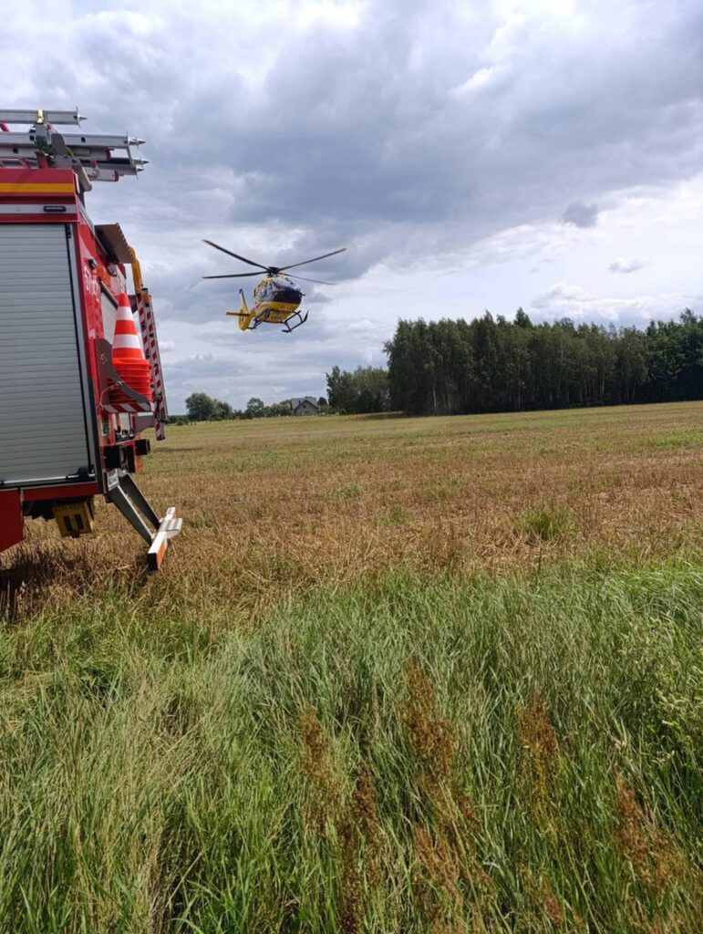 Śmigłowiec ratunkowy przelatuje nad polem, obok widać wóz strażacki z widocznymi sygnalizatorami.