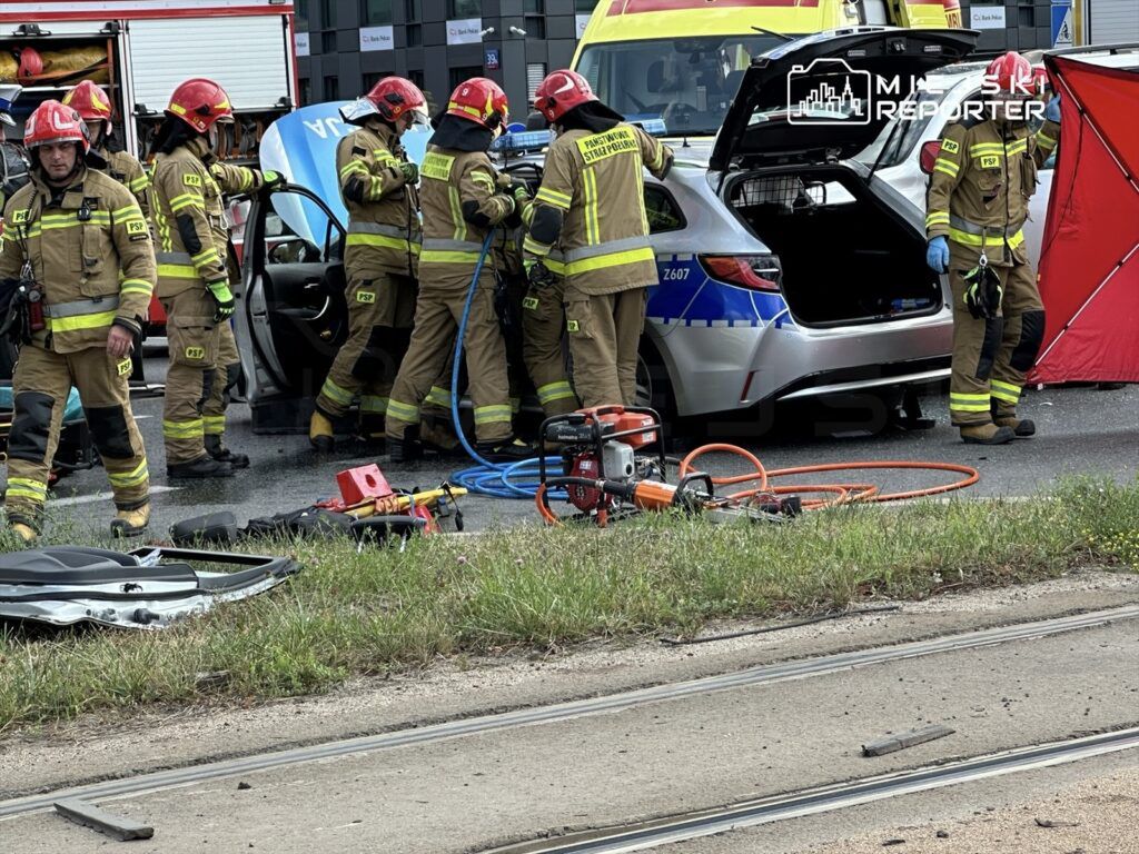 Grupa strażaków w mundurach i hełmach pracuje nad rozbitą osobówką, używając sprzętu ratunkowego na drodze.
