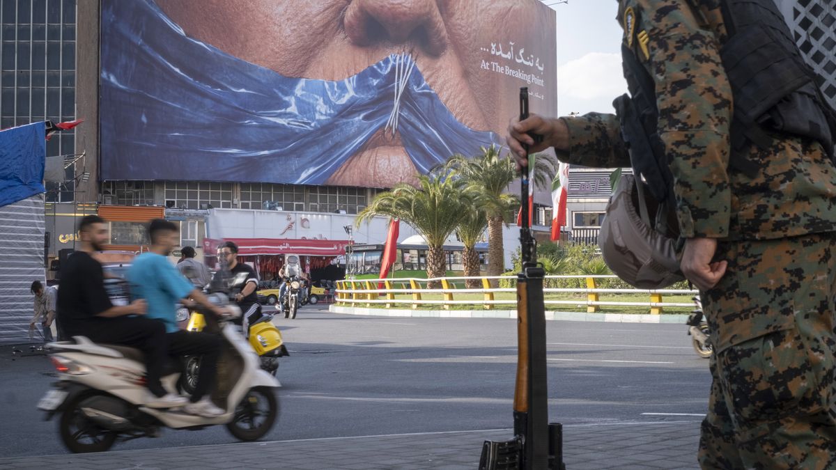 An armed Iranian police officer holding a rifle monitors the area as motorcyclists ride beneath a billboard depicting an AI-generated image of the Strait of Hormuz and an effigy of U.S. President Donald Trump, displayed on the wall of a state building in downtown Tehran, Iran, on May 3, 2026. (Photo by Morteza Nikoubazl/NurPhoto via Getty Images)