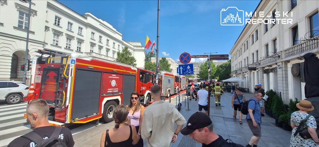 Grupa ludzi przechodzi obok dwóch wozów strażackich zaparkowanych na ulicy, w tle widoczne budynki i znaki drogowe.