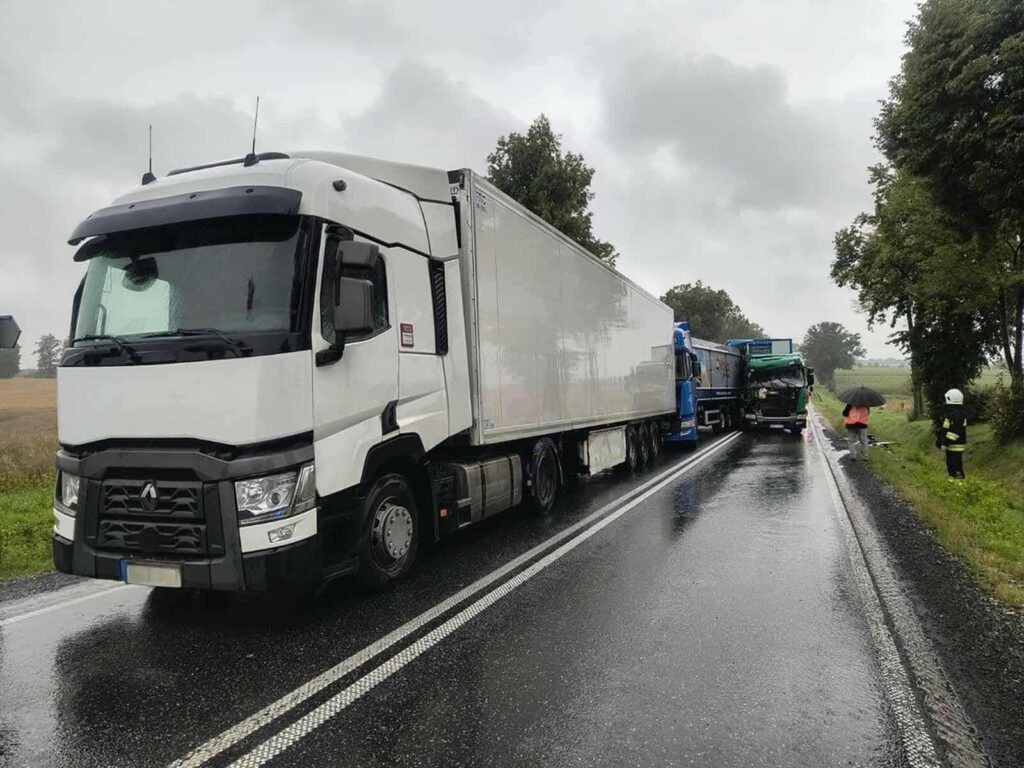 Czarny samochód ciężarowy z uszkodzoną kabiną stoi na drodze, obok niego pracują służby ratunkowe w deszczowej aurze.