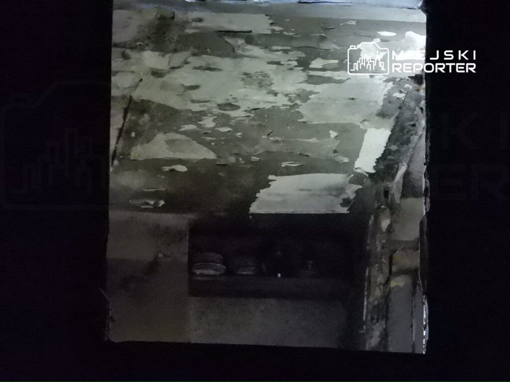 Słabo oświetlona, zniszczona ściana z łuszczącą się farbą i widocznym fragmentem półki z przedmiotami.