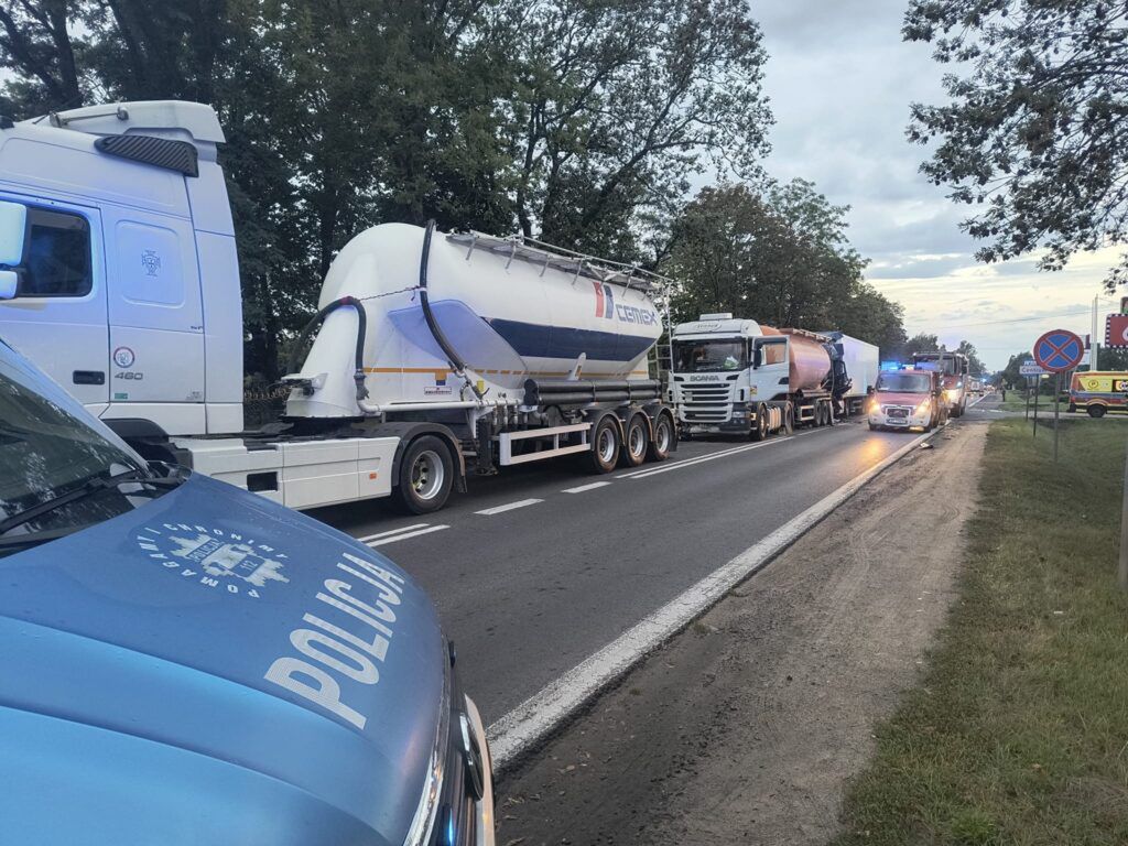 Kolumna ciężarówek z cysternami zatrzymana na drodze, obok policyjny radiowóz oraz włączone sygnały świetlne.