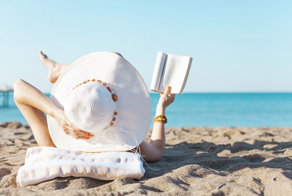 kobieta opala się na plaży w kapeluszu i czyta książkę