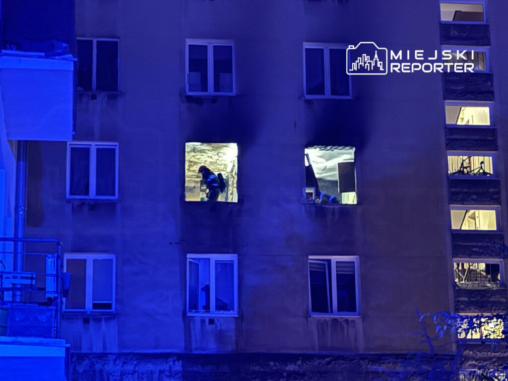 Strażak w sprzęcie ochronnym przeszukuje zadymione mieszkanie przez otwarte okno w bloku mieszkalnym.