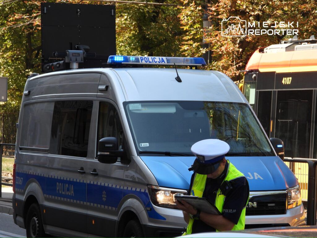 Policjant w kamizelce odblaskowej spisuje notatki przed policyjnym pojazdem z włączonymi sygnałami świetlnymi.