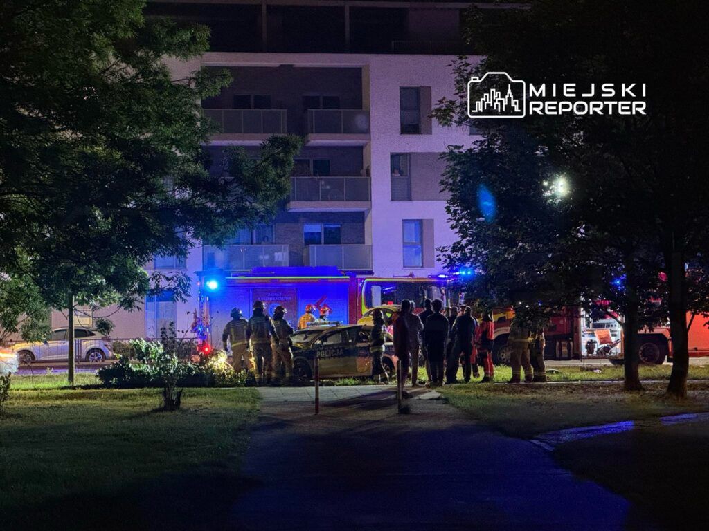 Grupa strażaków i ratowników medycznych udziela pomocy przy wypadku drogowym w nocy, w pobliżu nowoczesnego budynku mieszkalnego.