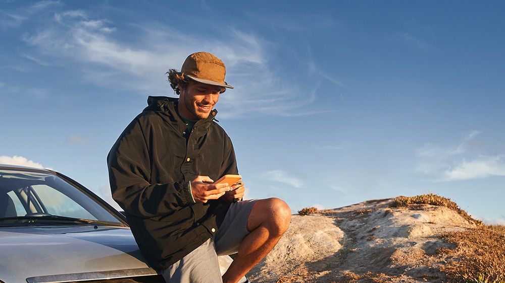  Mężczyzna siedzący na masce samochodu, uśmiechając się i trzymając telefon w rękach, na tle błękitnego nieba
