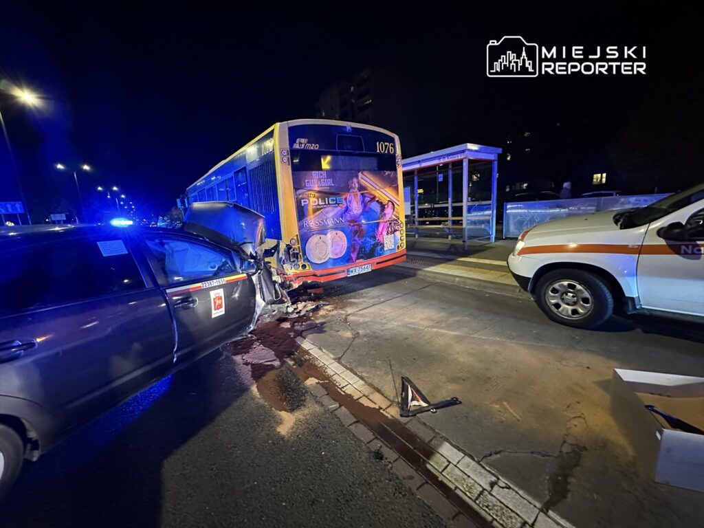 Czarny samochód osobowy z uszkodzeniami zderza się z autobusem na oświetlonej ulicy nocą, obok stoi pojazd służb ratunkowych.