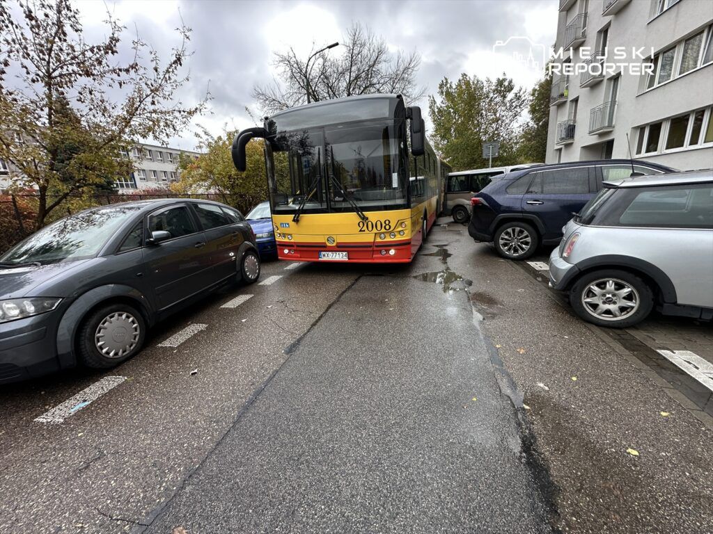 Autobus komunikacji miejskiej zatrzymany na wąskiej ulicy, otoczony przez zaparkowane samochody osobowe.