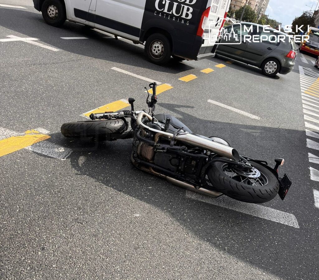 Motocykl leżący na jezdni, na skrzyżowaniu ulic. W tle widoczne pojazdy dostawcze oraz osobowe. Sytuacja sugeruje możliwy wypadek lub awarię jednośladu. Na drodze widoczne oznaczenia poziome. Zdjęcie dokumentuje zdarzenie drogowe w miejskim otoczeniu.