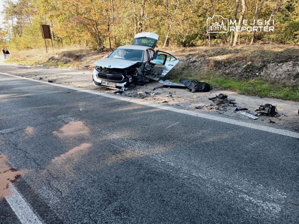 Zniszczony samochód osobowy stoi na poboczu drogi, obok leżą fragmenty wraku i plamy oleju. W tle widoczna zieleń drzew.