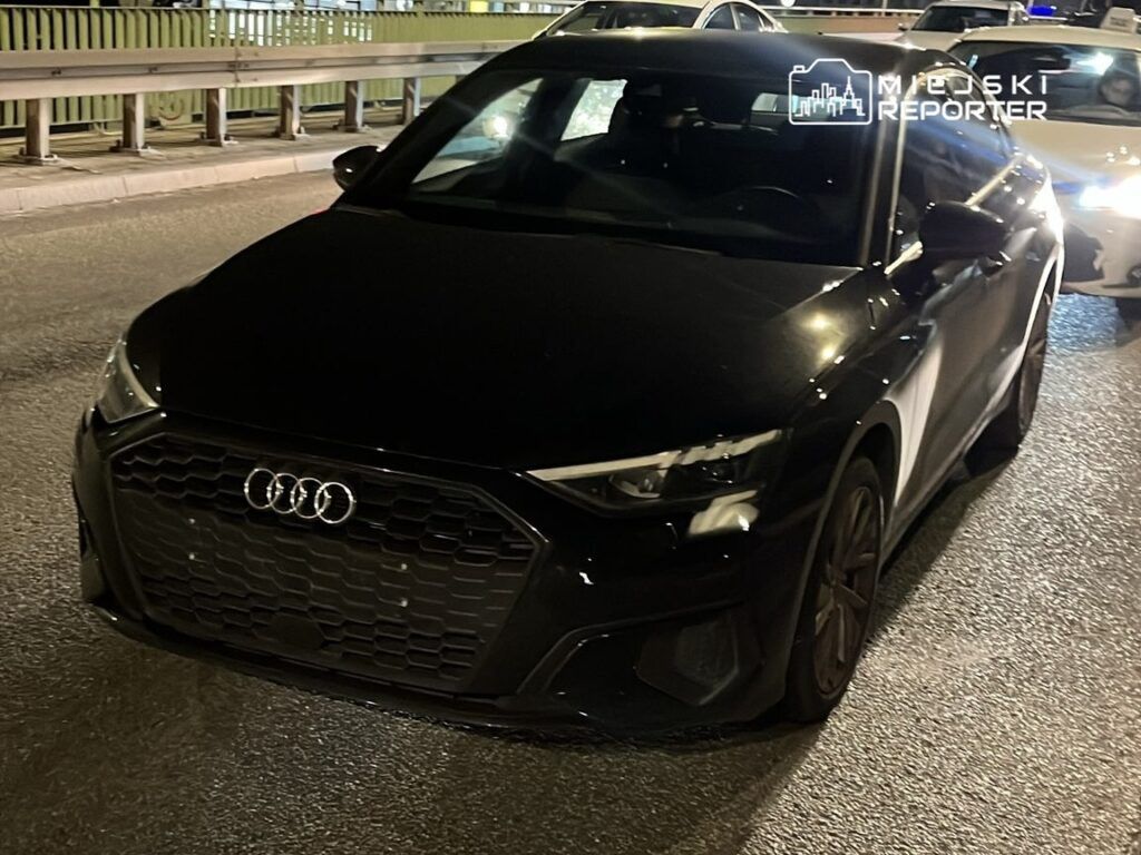 Czarny samochód marki Audi zaparkowany na oświetlonej ulicy w nocy, w otoczeniu innych pojazdów.