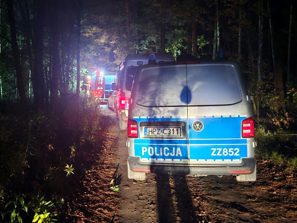 Policyjny van zaparkowany na leśnej drodze, w tle widoczne światła wozów strażackich.